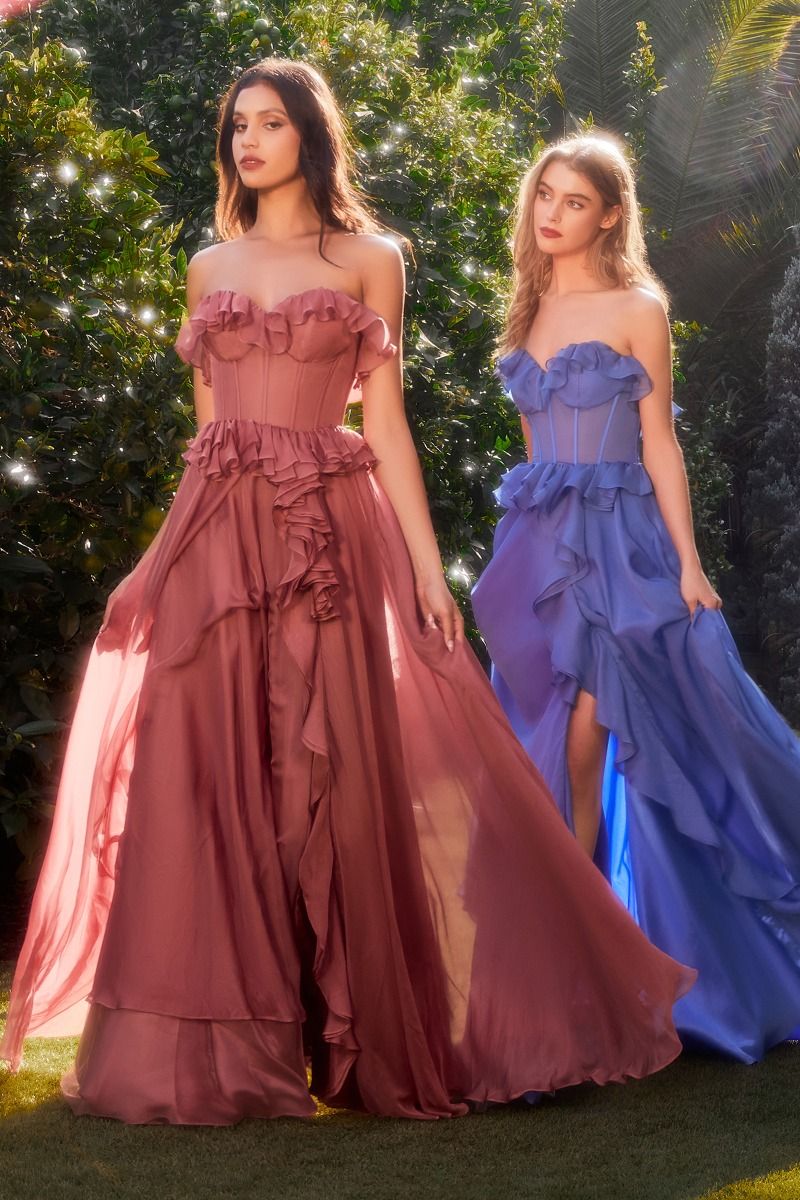 Two women stand outdoors in sunlight, each wearing the Andrea and Leo A1341 Strapless Ruffled Prom Dress—one in mauve pink, one in blue—creating a stunning contrast with the lush green foliage.